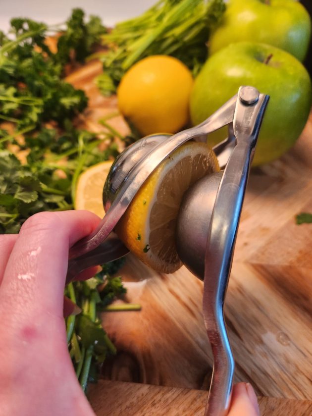 hand squeezing a lemon with a mini juicer. Green apples, lemon and parsley in the background