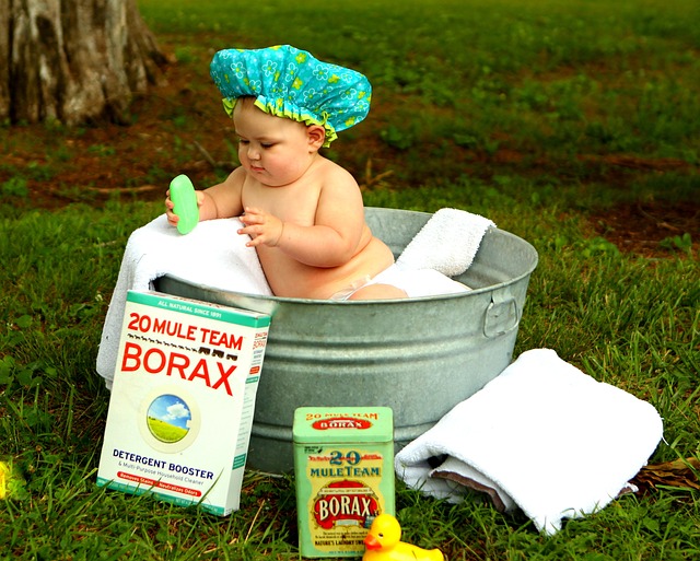 white baby sitting in a tin tub with a shower cap on head and borax and cleaning supplies around the tub