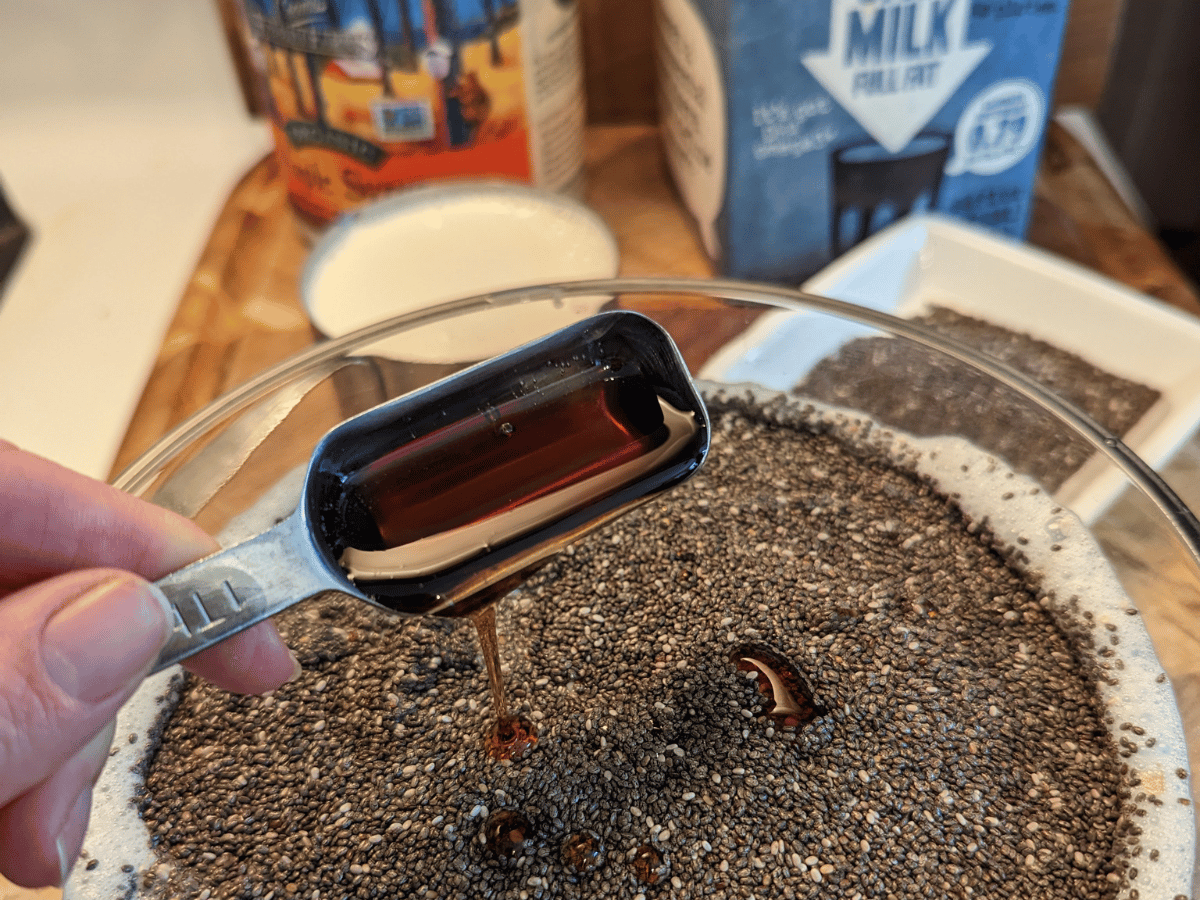 glass bowl with milk and chia seeds. A tablespoon of maple syrup being poured into the bowl. Oat milk and maple syrup containers in the background