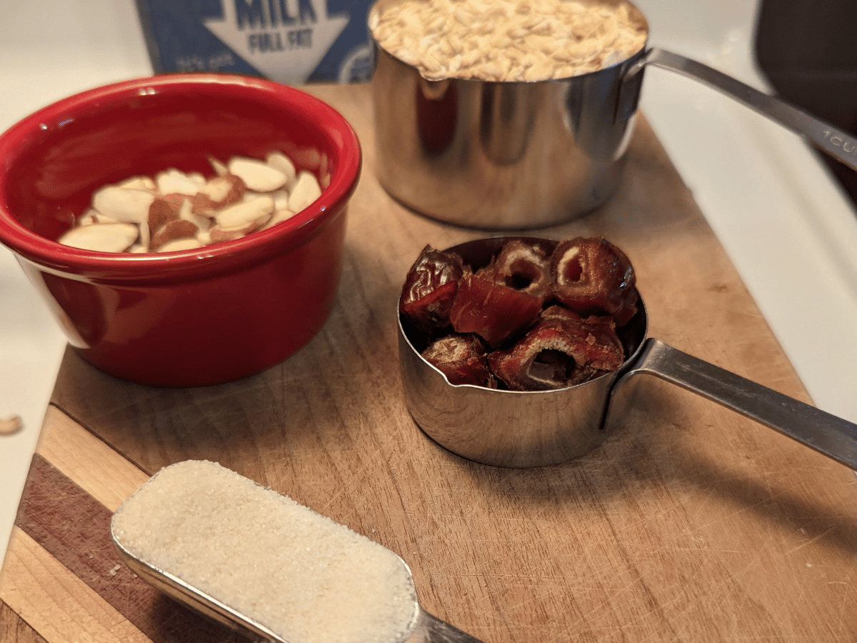 Wood cutting board with a tbsp of sugar, 1/4 cup of dried dates, 1 cup of dry oats, a small red bowl of sliced almonds, and a carton of oat milk in the background