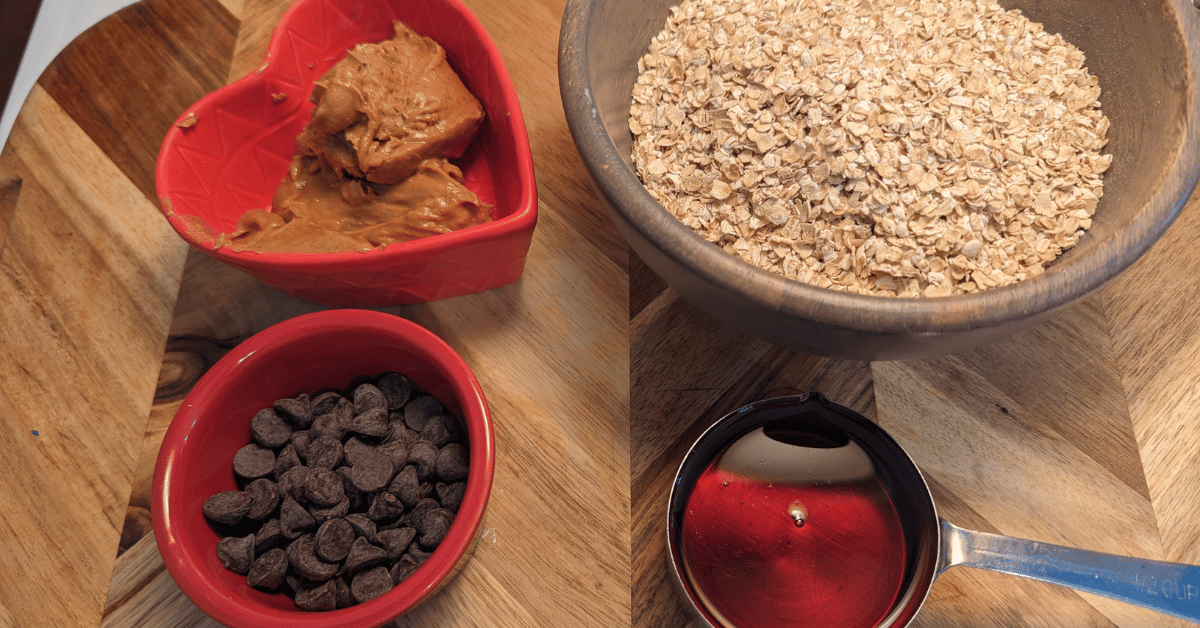 one red bowl with peanut butter, one red dish with chocolate chips, a wood bowl with dry oats, and 1/4 cup with maple syrup, all on a wood cutting board